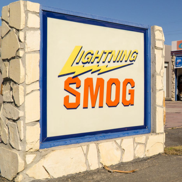 Blue-framed roadside sign for Lightning Smog on a stone wall, with yellow and orange lettering.